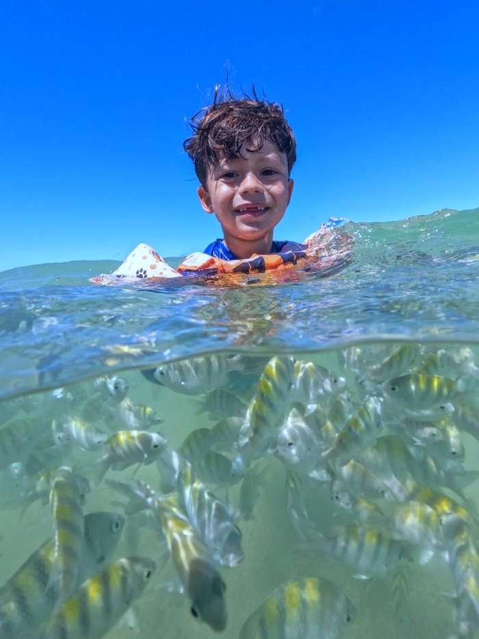 Criança sorrindo na água das Piscinas Naturais do Seixas cercada de peixinhos
